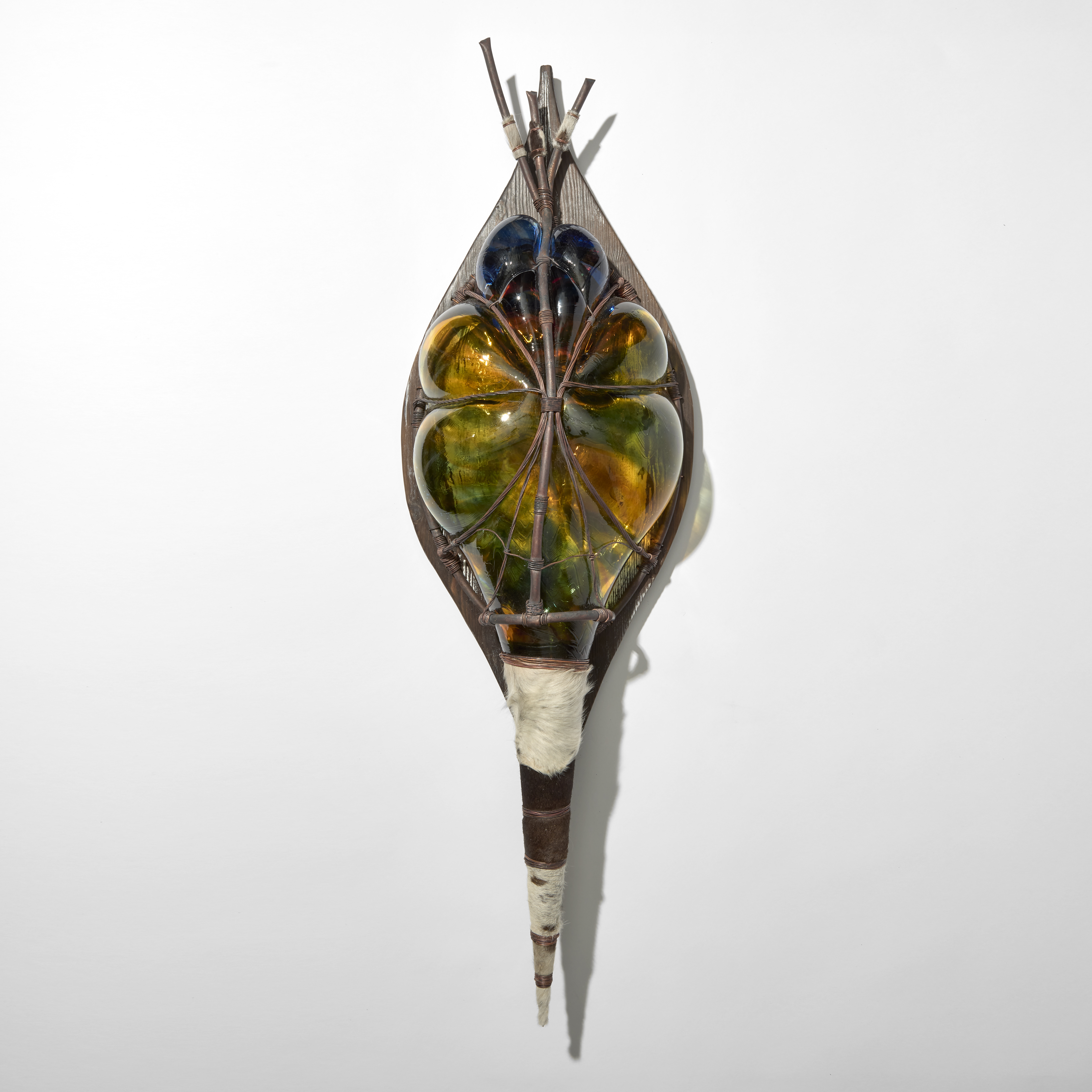 stylised african wall mounted shield created from amber blue and green glass blown through a copper cage mounted on a wooden back board and with a pointed base wrapped in cow hide