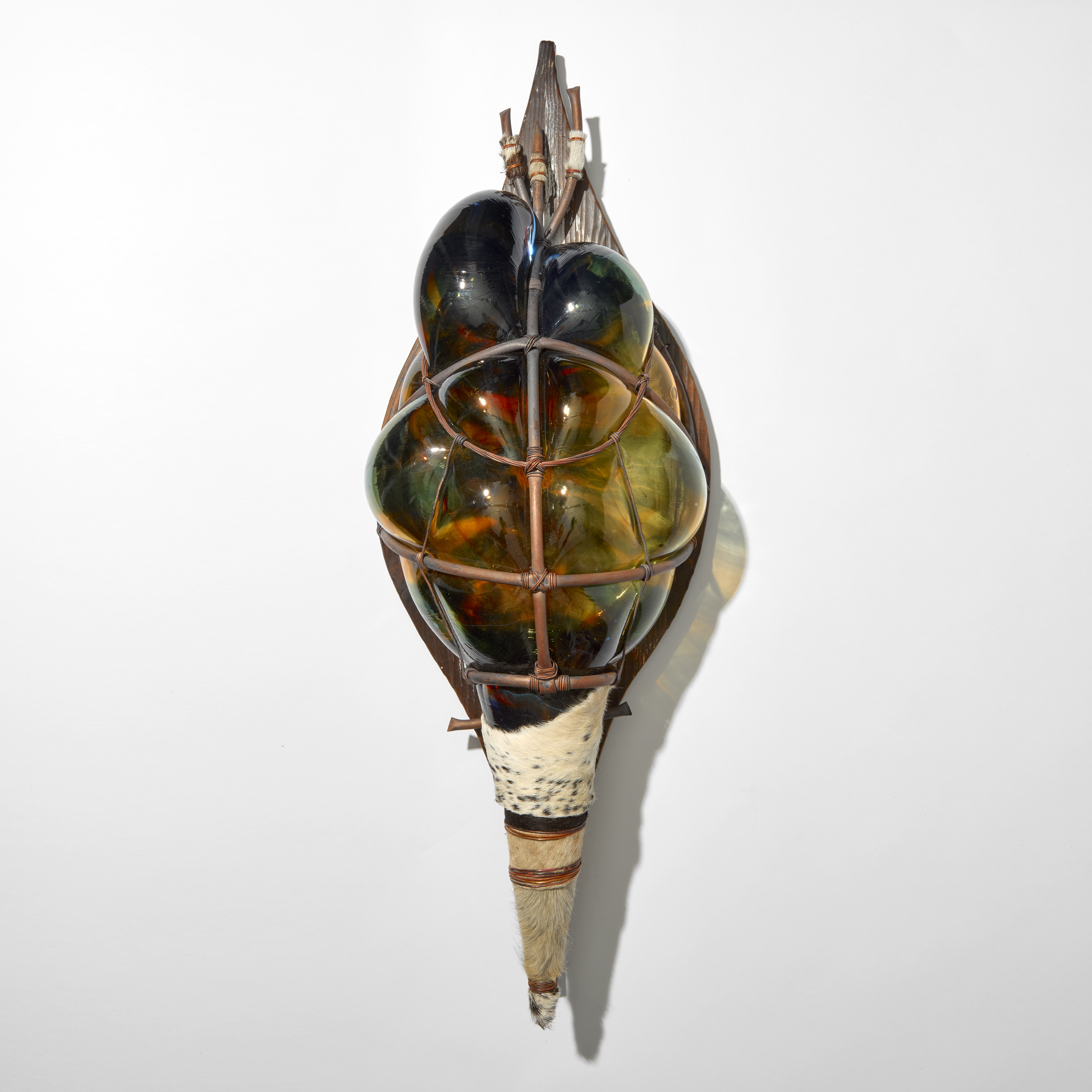 african stylised shield made from a cage created from copper pipe with blown glass in blue green and amber bulging through the gaps strapped with a wooden back board and with a pointed base wrapped in cow hide