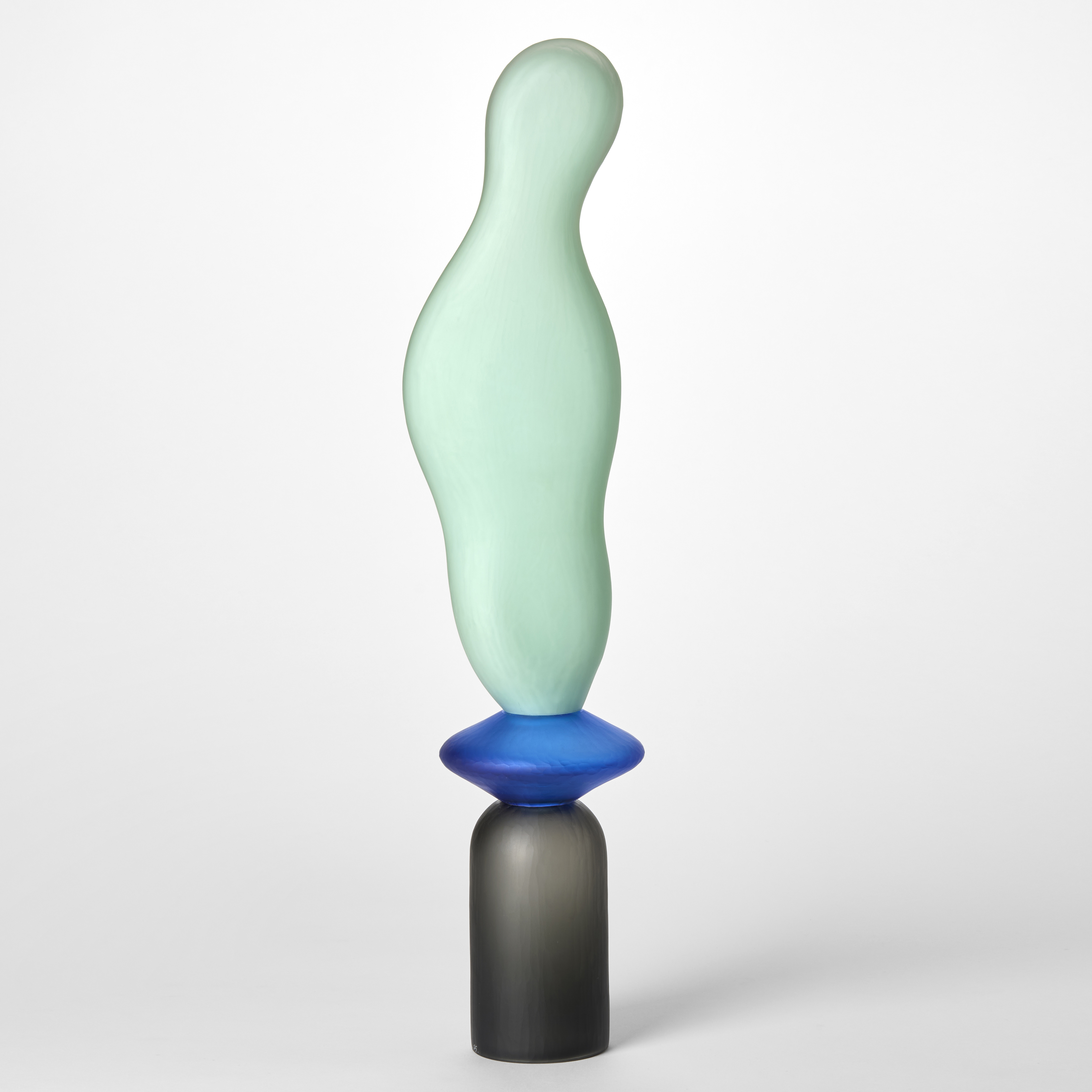 tall opaque sculpture with a textured surface created from three stacked shapes a grey upside down cup a blue disc and a mint long undulating balloon hand blown from glass