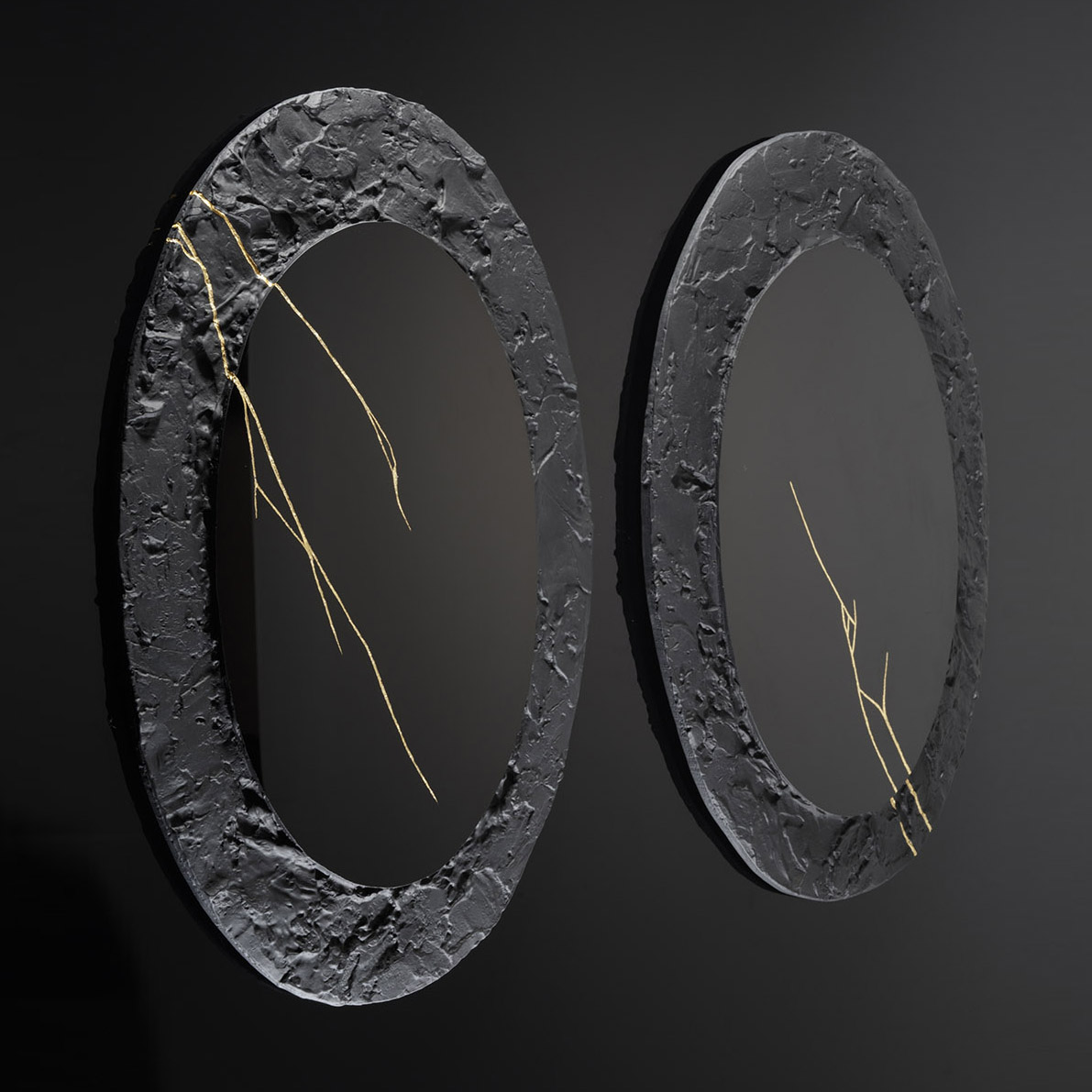 two black wall hung oval discs with thick roughly textured borders and cracks across the surface in gold with central polished black mirrors