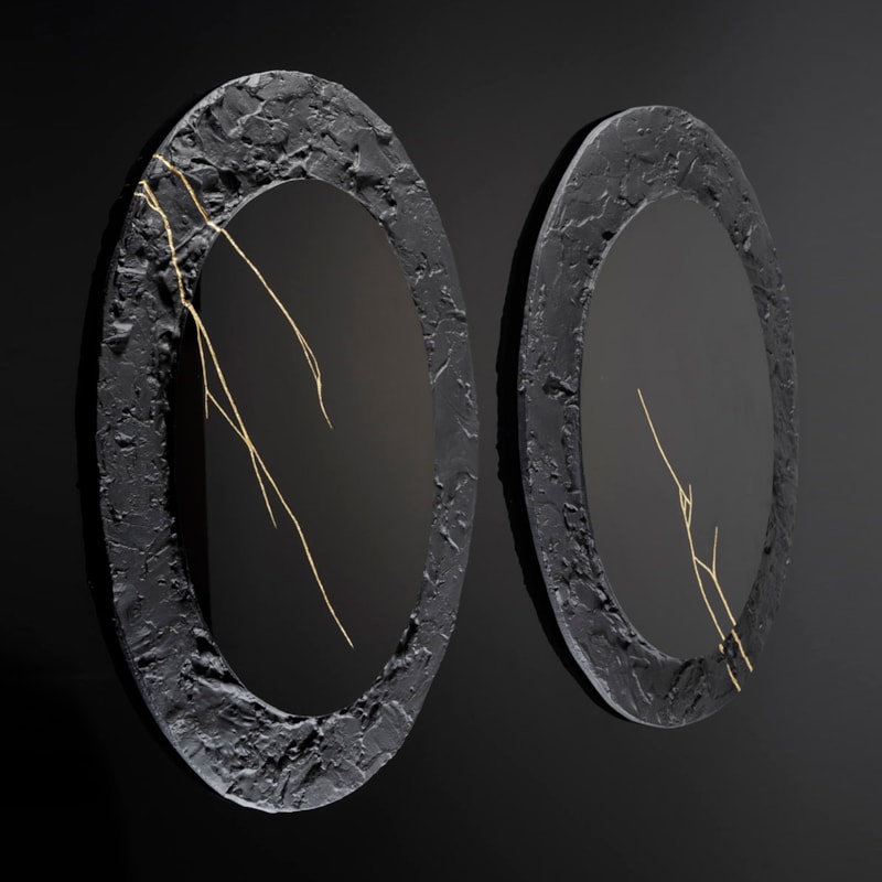 two black wall hung oval discs with thick roughly textured borders and cracks across the surface in gold with central polished black mirrors