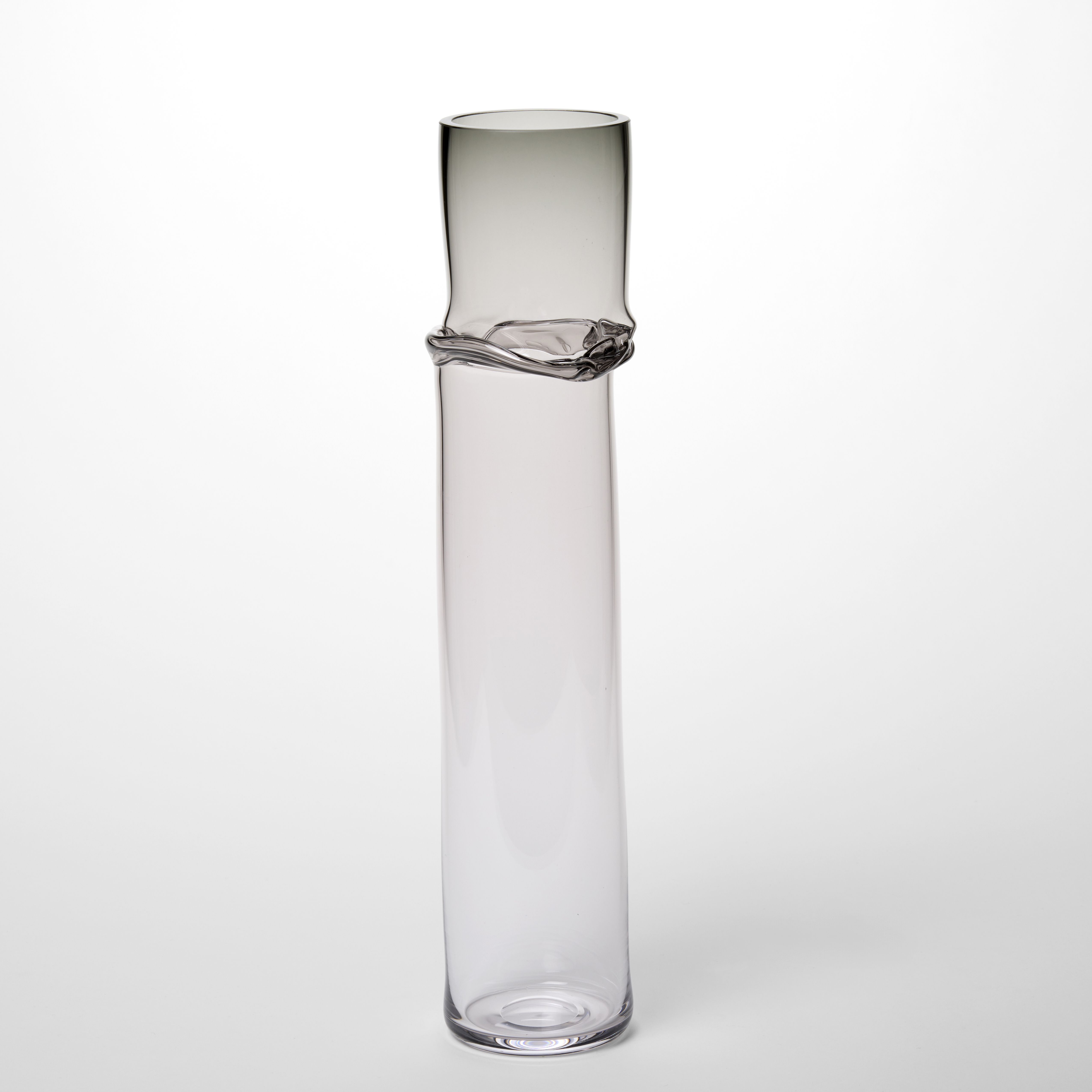 tall cylindrical glass sculpture with a minimalist design that is mostly clear with a subtle gradient from transparent at the base to smoky grey at the top and around the upper section it has a delicate uneven ridge encircling the form adding a natural organic detail to the otherwise smooth and polished surface hand made from blown glass
