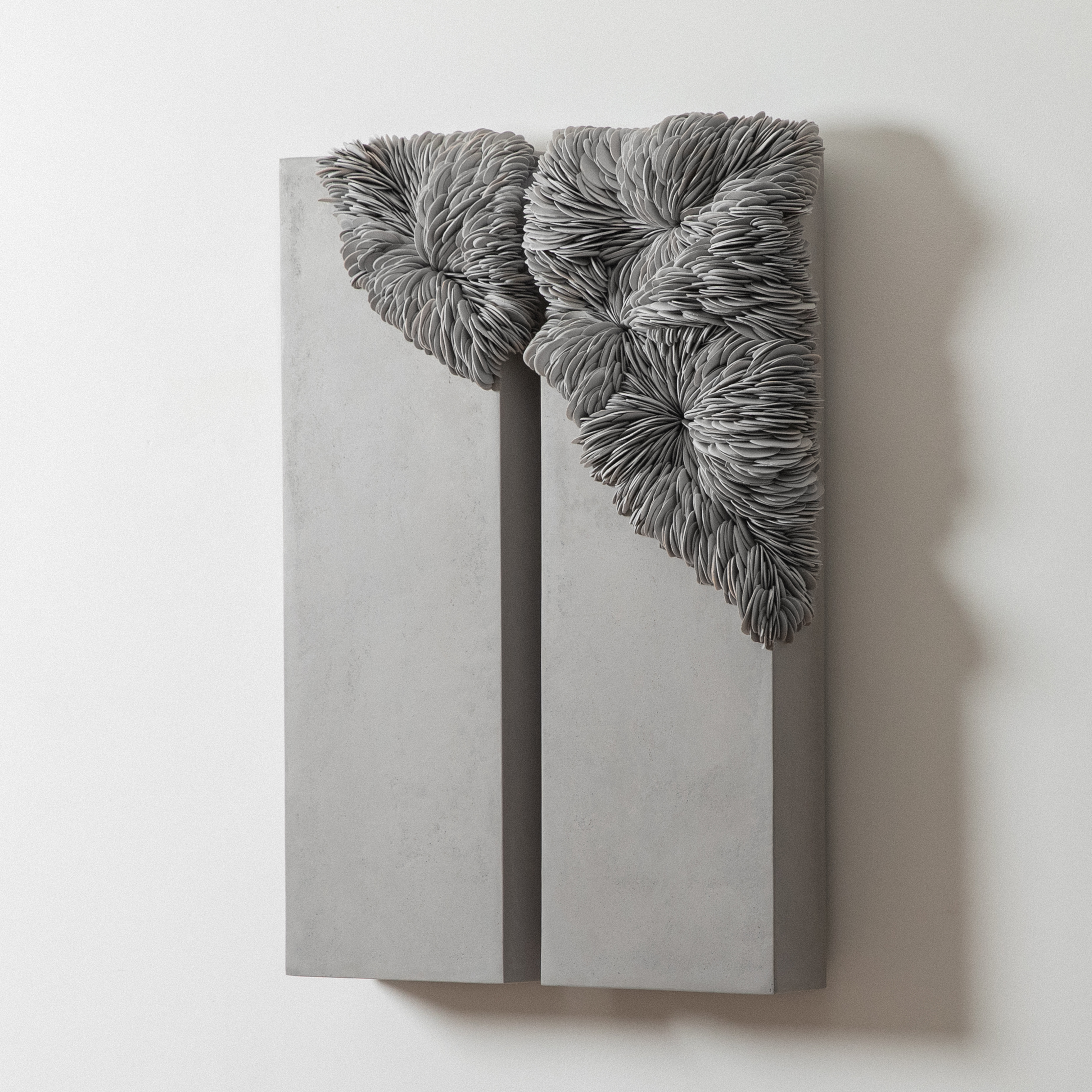 wall mounted organic artwork consisting of two identical long chunky grey panels with the upper sections featuring a relief pattern of curved interlocking shards traversing from one panel to the next hand made from porcelain and tadelakt plaster