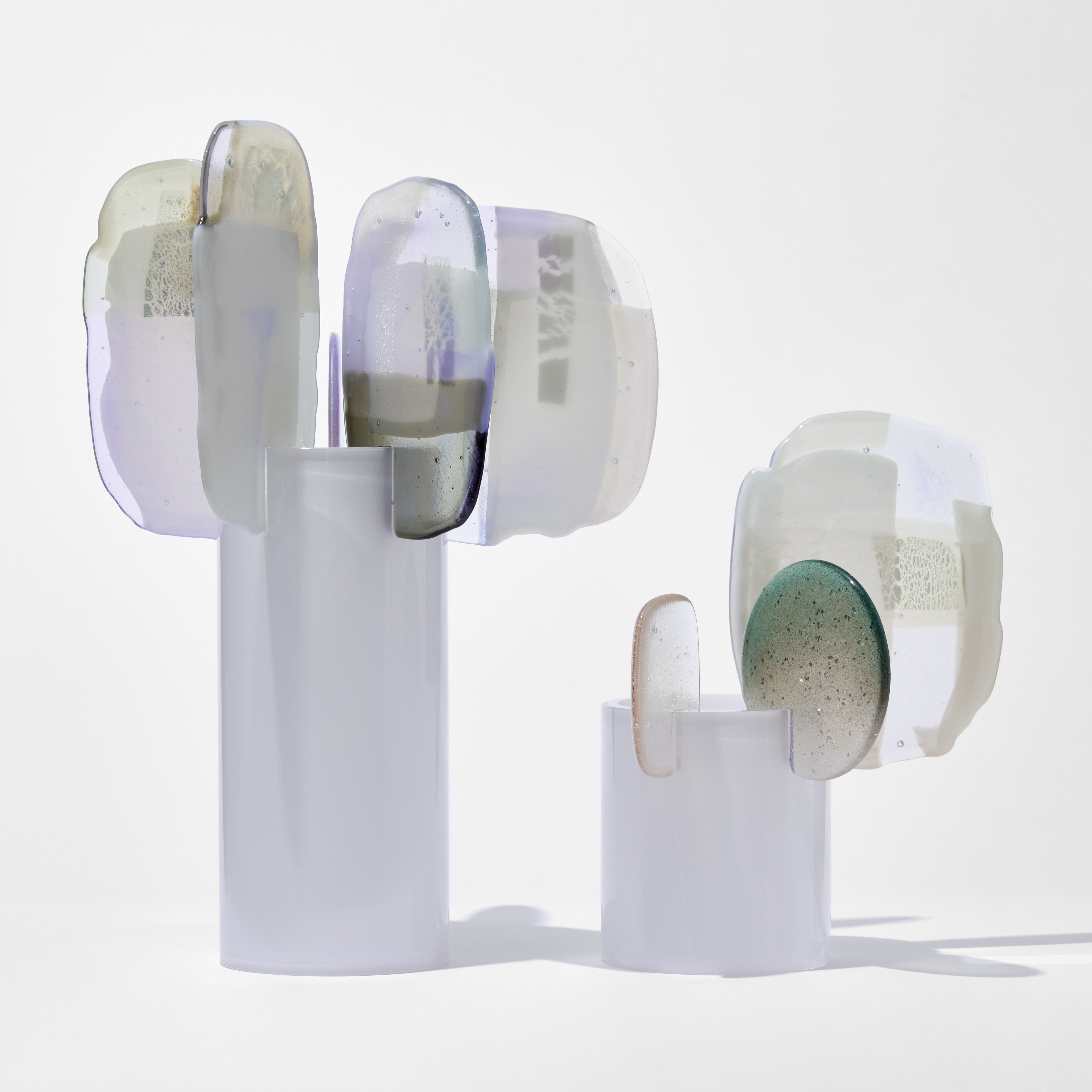 two soft opaque white standing forms with glossy cylinder trunks with organic flat finials perched on the tops edges with mixed abstract patterns on the surface  in grey alabaster teal and lilac hand made from blown and fused glass