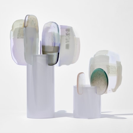 two soft opaque white standing forms with glossy cylinder trunks with organic flat finials perched on the tops edges with mixed abstract patterns on the surface  in grey alabaster teal and lilac hand made from blown and fused glass