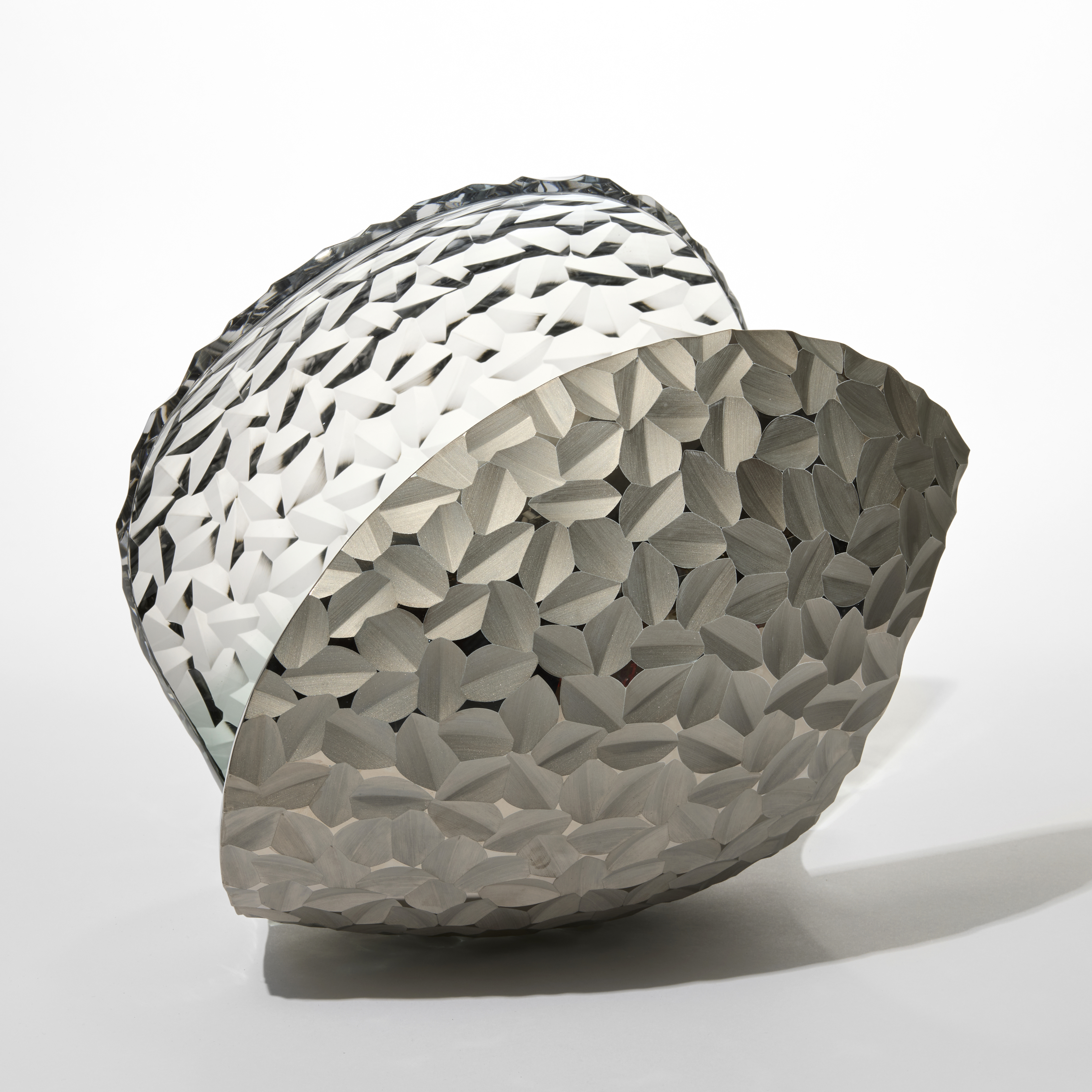 curled simplified shell like shape with the surface covered in repeat incised tessellated pattern handblown from light grey glass with a one side covered in matt silver lustre