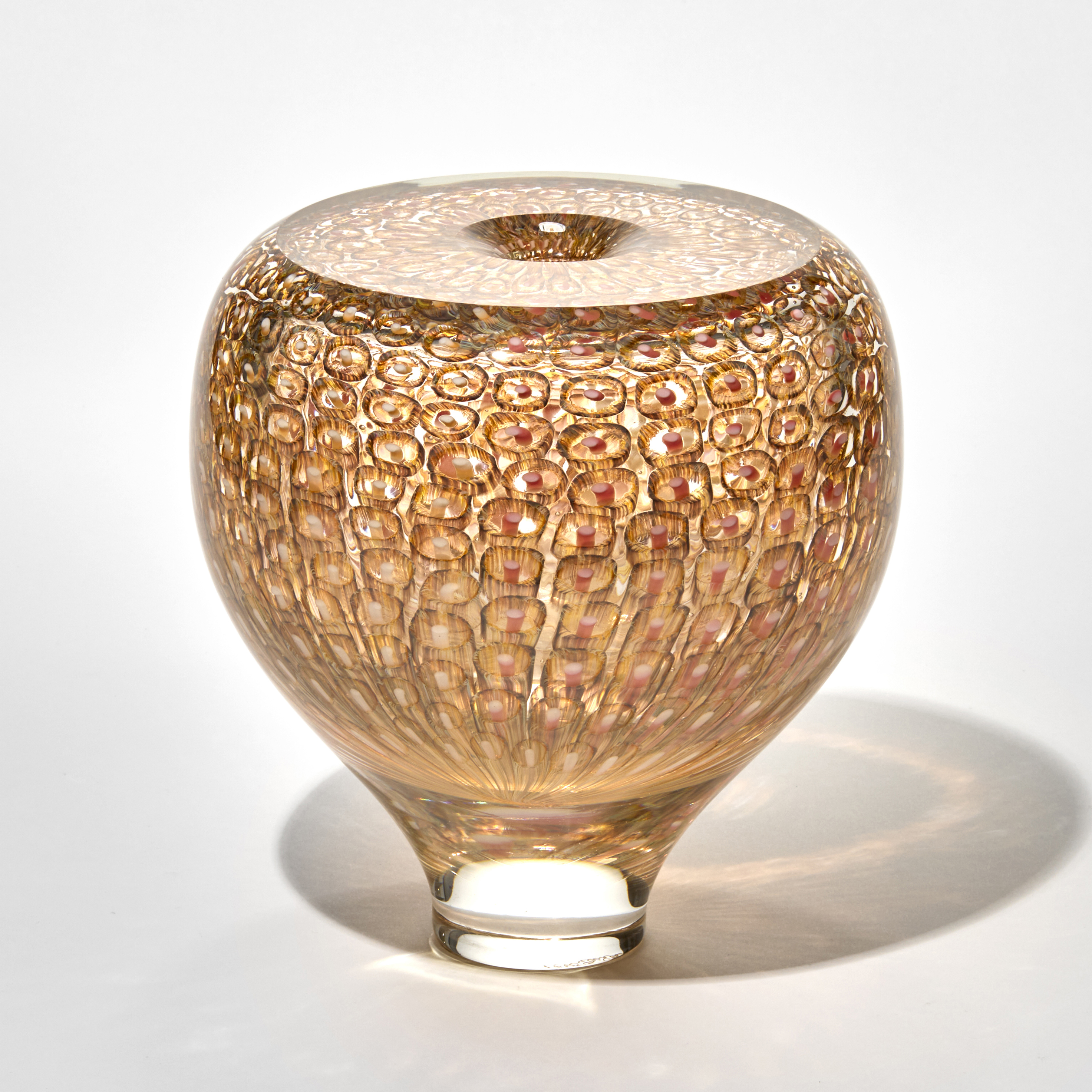 rounded vase like sculpture in warm amber tones with a narrow base and wide body with the surface covered in a honeycomb like murrini patterns of circular impressions creating a textured reflective effect and a top a small central opening hand made from blown glass