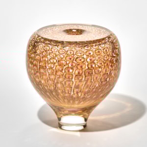rounded vase like sculpture in warm amber tones with a narrow base and wide body with the surface covered in a honeycomb like murrini patterns of circular impressions creating a textured reflective effect and a top a small central opening hand made from blown glass
