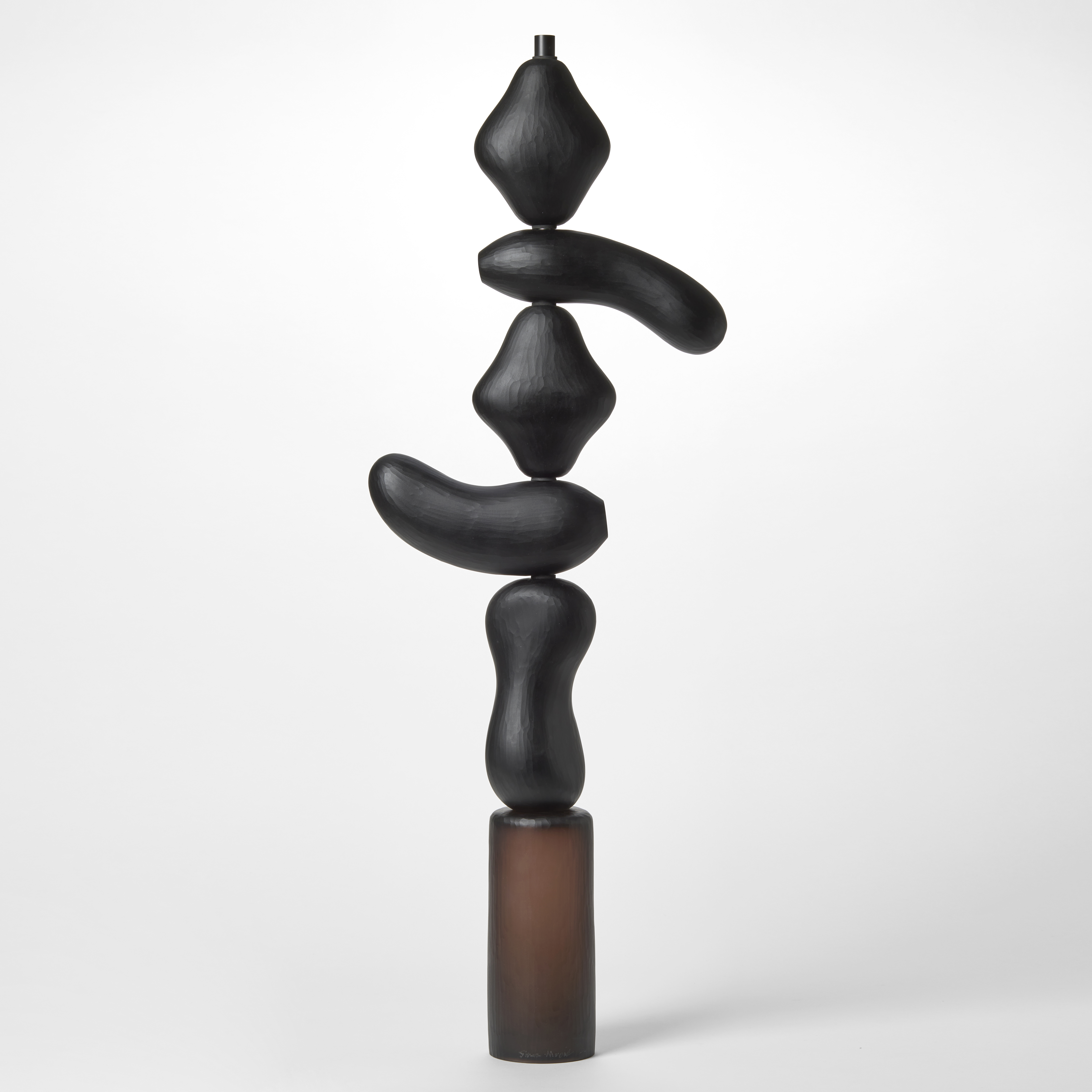 opaque abstract tall totem sculpture consisting of five geometric stacked shapes in black perched on a cylindrical brown base made from hand blown glass with a repeat surface texture