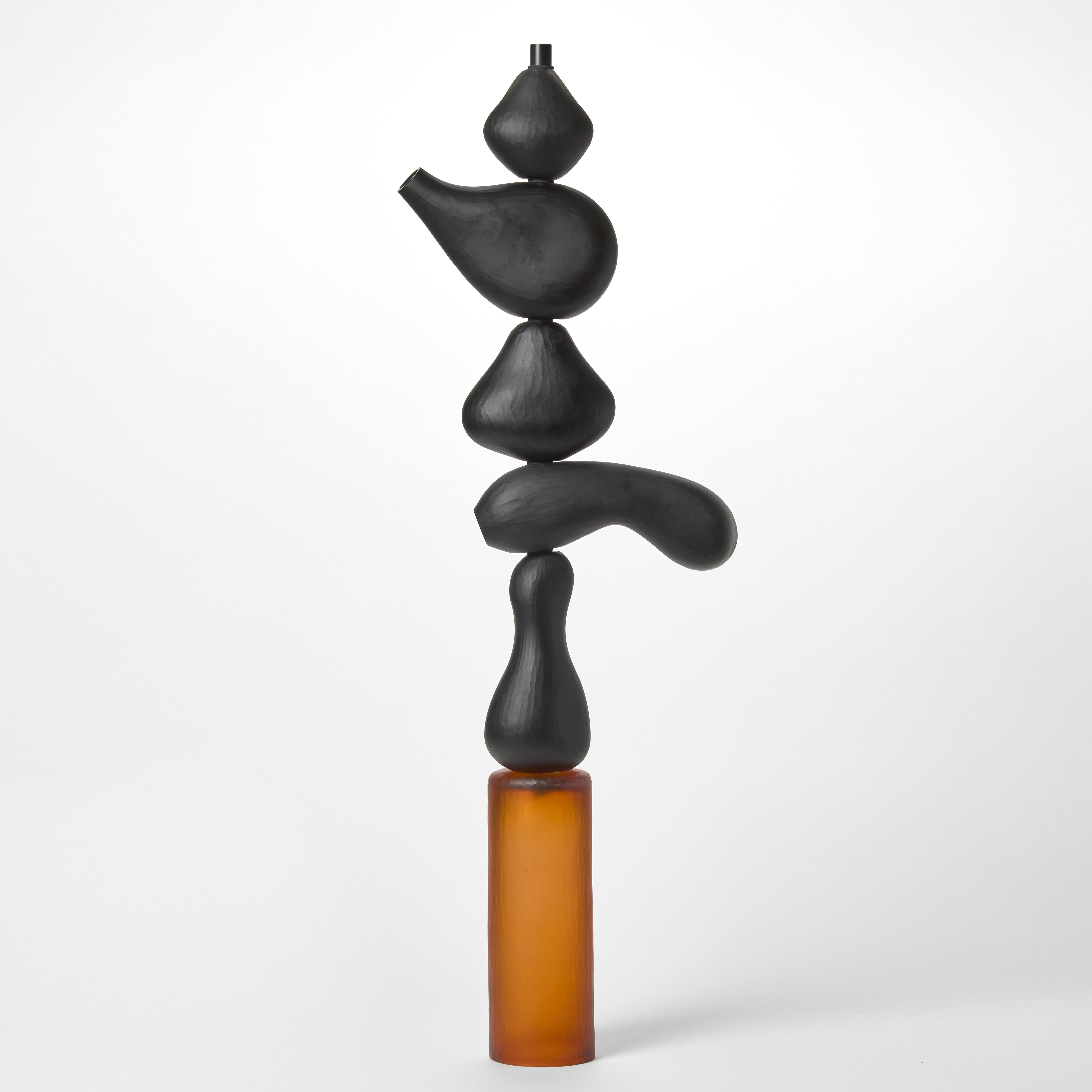 tall totem sculpture consisting of six stacked shapes five in opaque black and the bottom one a burnt orange cylinder hand made from blown glass with a repeat shallow surface texture