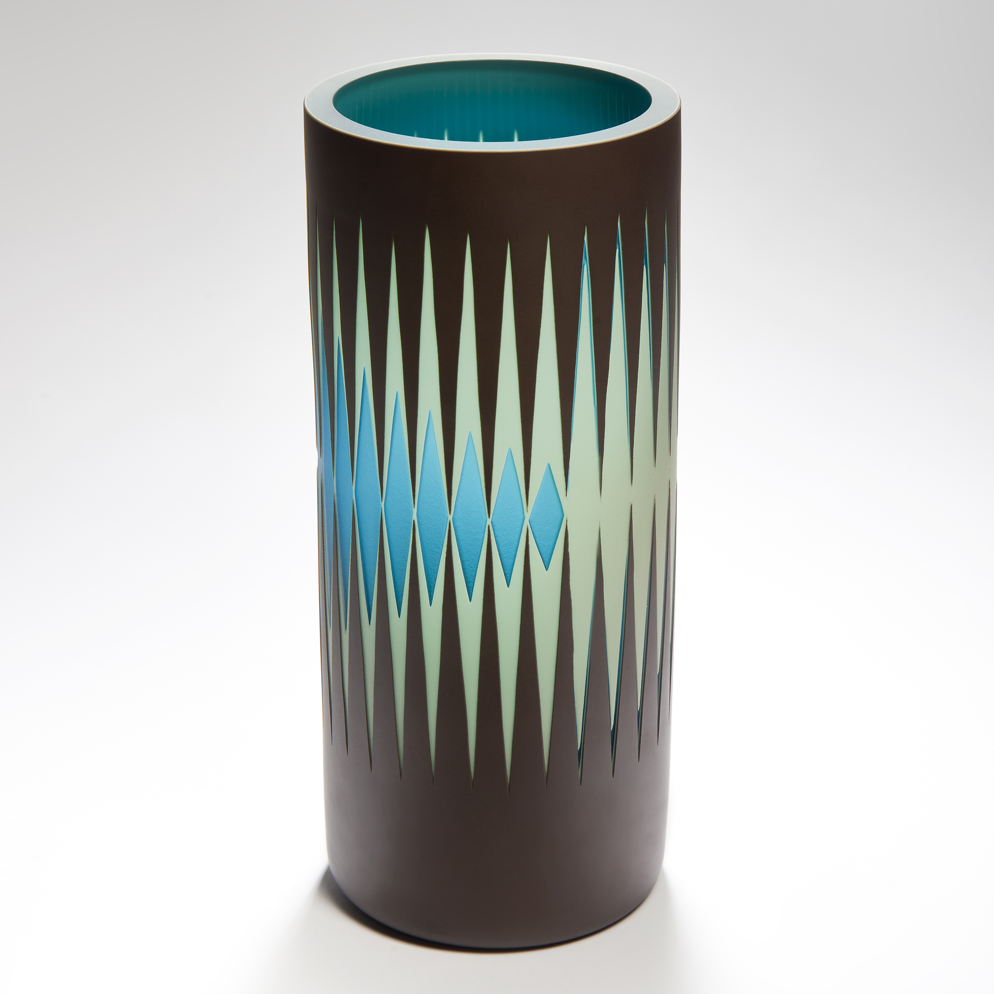 tall cylindrical vase with a reverse diminishing geometric pattern of repeat stretched diamonds and pointed spikes in light jade green and turquoise with a black background handmade from glass