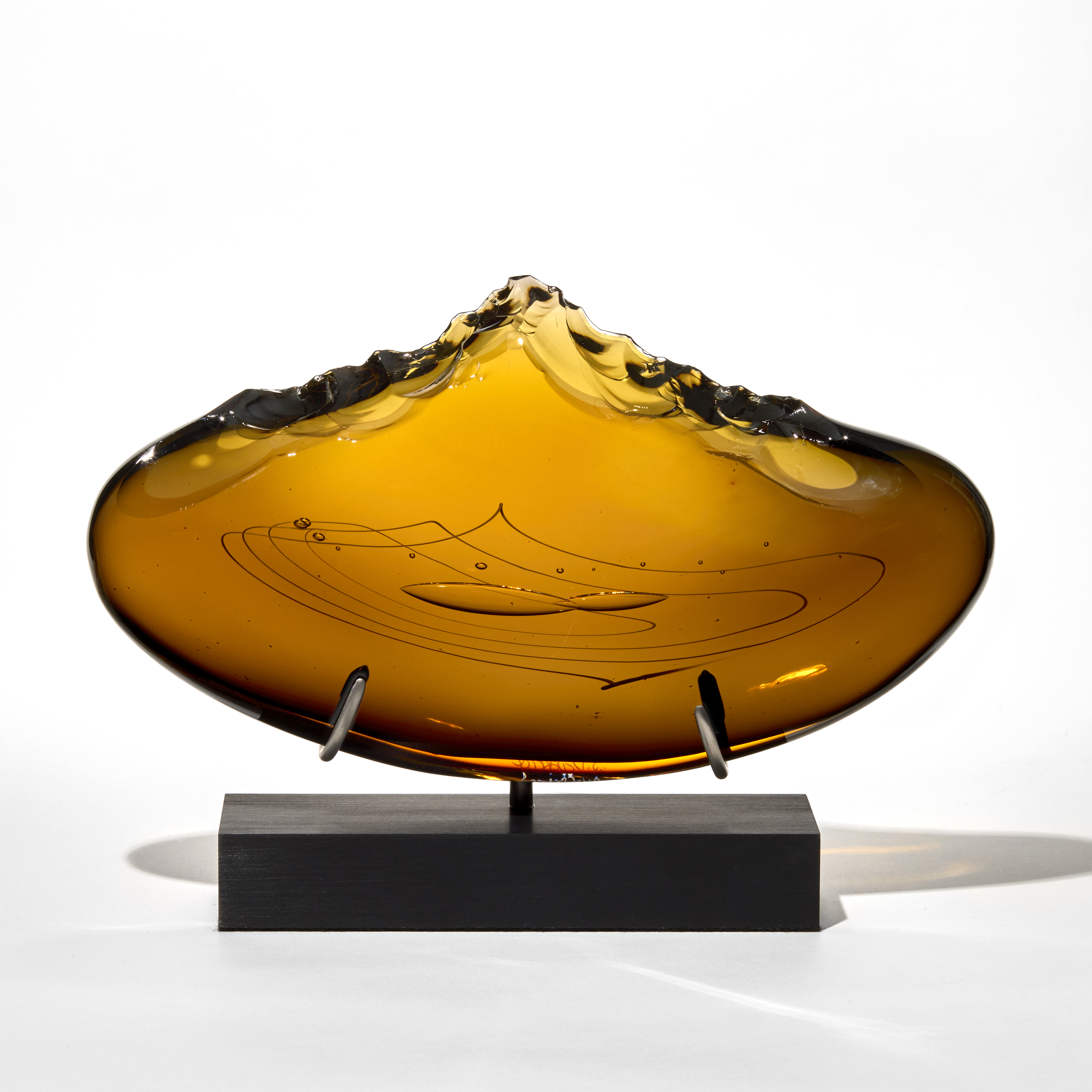 rich amber coloured sculpture with a wide curved and flattened oval form featuring smooth surfaces and line patterns and bubbles trapped in the centre with a top irregular and beaten edge resembling the stone tools made by early man mounted upright on a sleek black rectangular metal base with two supports hand blown from glass