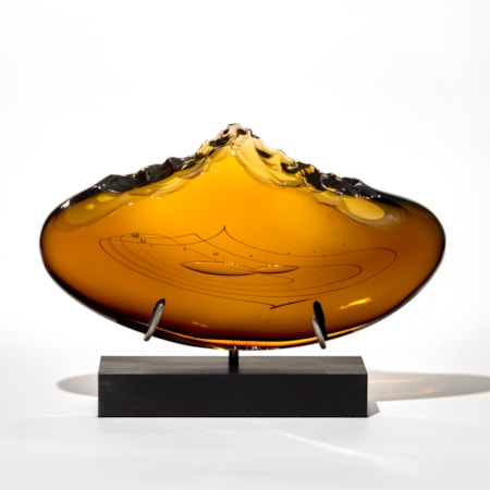 rich amber coloured sculpture with a wide curved and flattened oval form featuring smooth surfaces and line patterns and bubbles trapped in the centre with a top irregular and beaten edge resembling the stone tools made by early man mounted upright on a sleek black rectangular metal base with two supports hand blown from glass