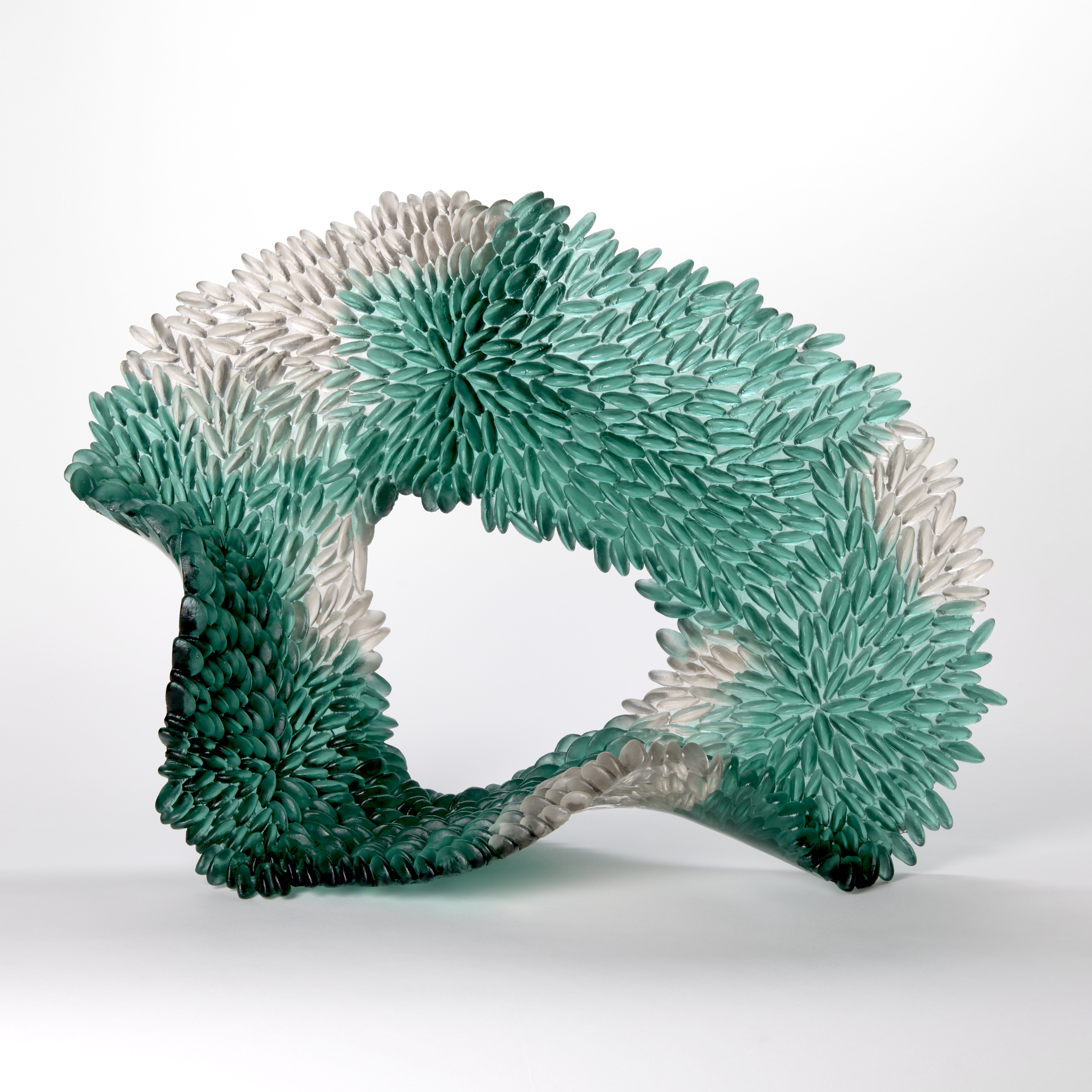 standing looped abstract sculpture in opaque teal green with patches of grey with the front surface covered in small repeat raised discs like scales hand made from cast and sculpted glass