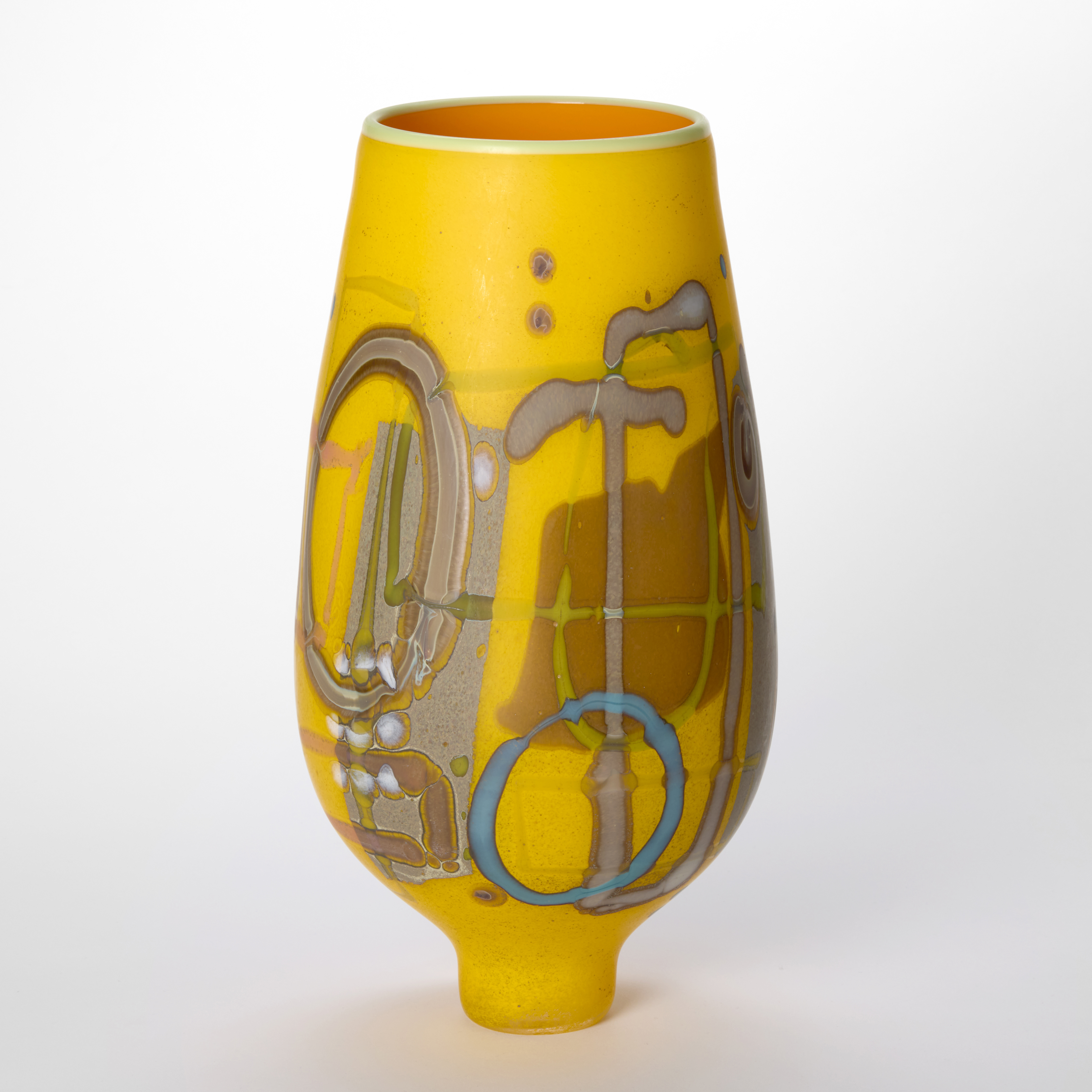 Bright rich matt yellow bulbous vase perched upon an elegant narrow foot with abstract surface patterns and motifs in brown fawn beige white and teal with a wide top opening and glossy interior in orange hand blown from glass