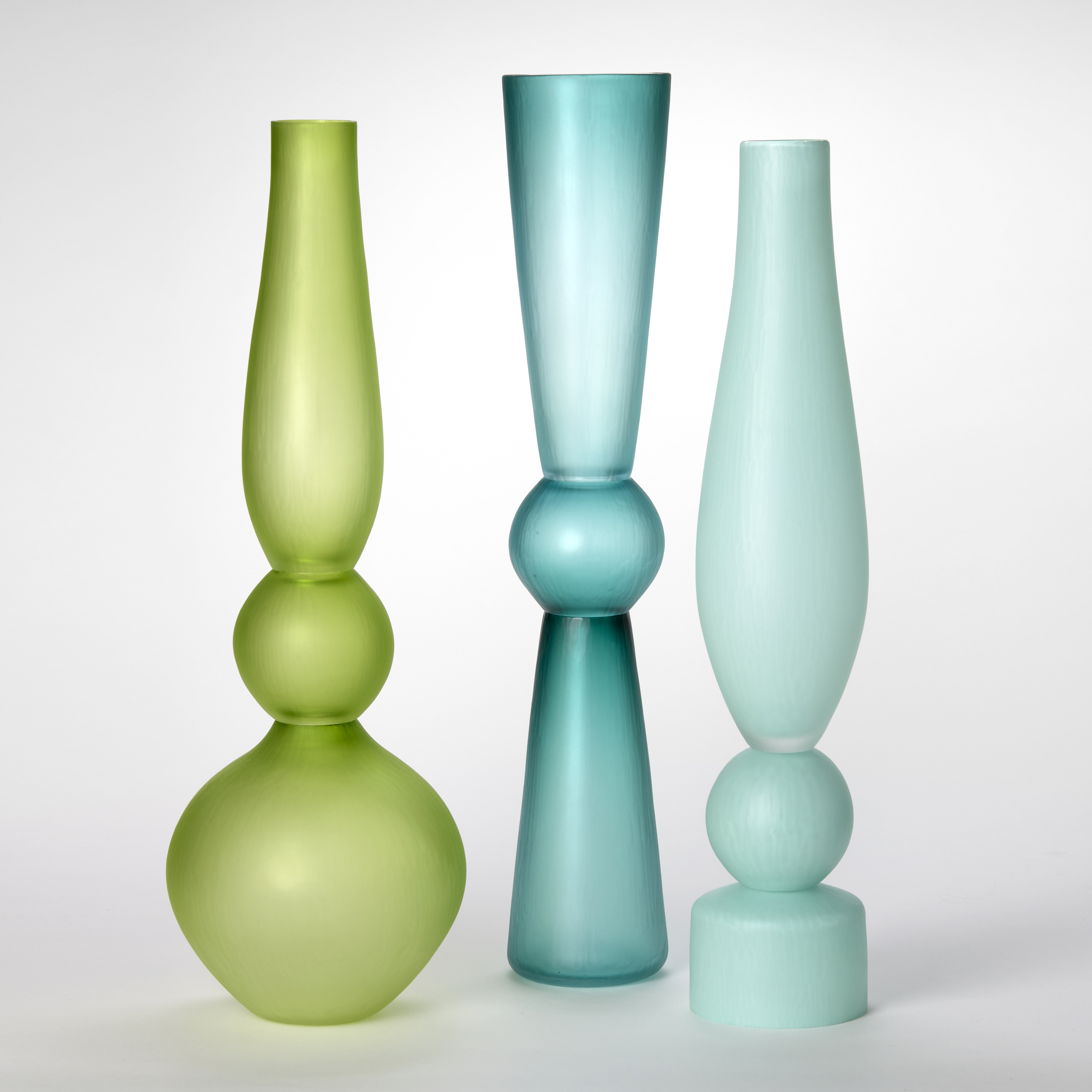 a set of opaque architectural tall standing vases each made of three stacked components and shapes and a soft satin surface finish with one in lime one in teal one in light blue and one in light green made from hand blown and cut glass