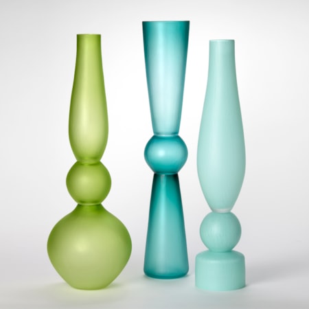 a set of opaque architectural tall standing vases each made of three stacked components and shapes and a soft satin surface finish with one in lime one in teal one in light blue and one in light green made from hand blown and cut glass