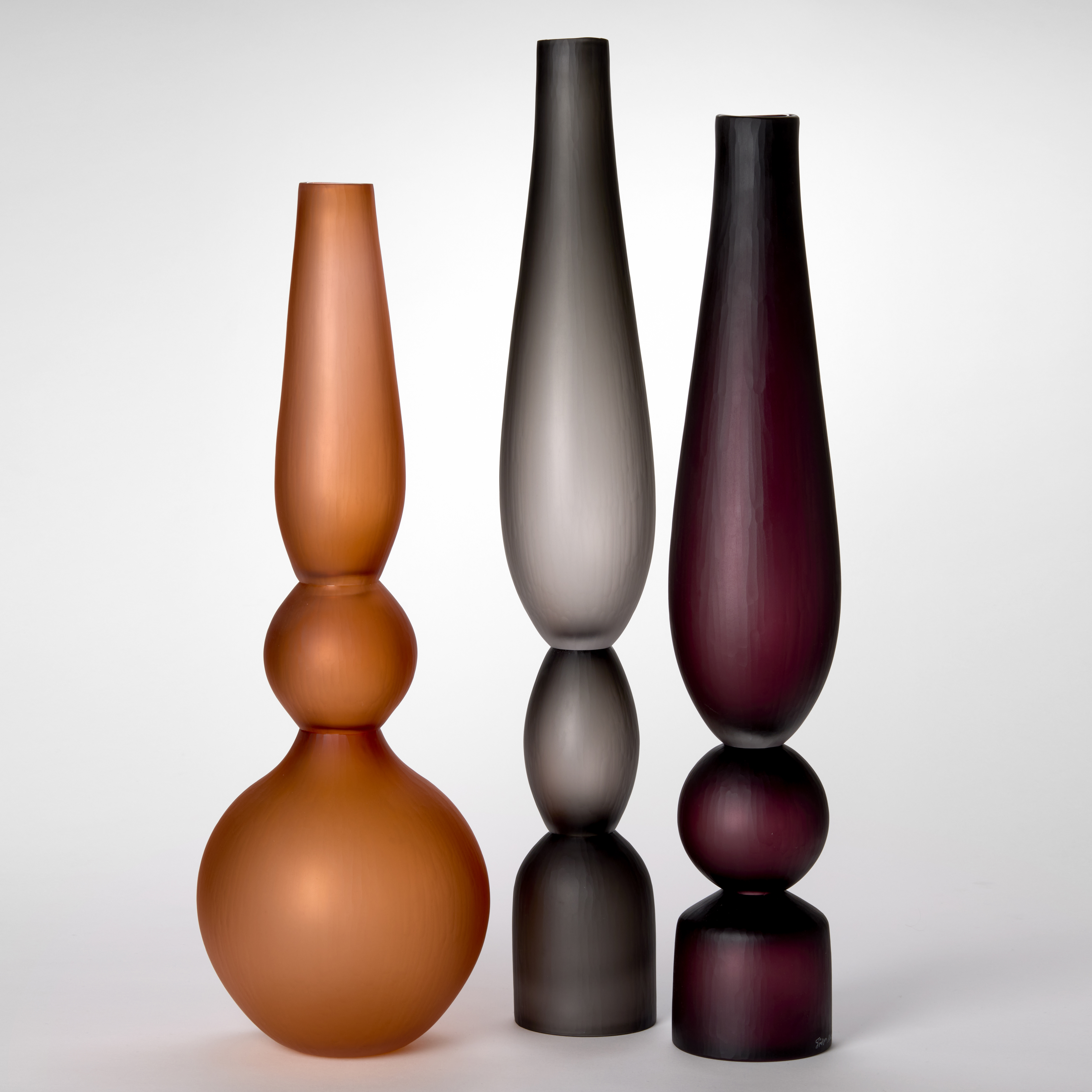trio of opaque architectural tall standing vases each made of three stacked components and shapes and a soft satin surface finish with one in turquoise one in grey and one in sienna made from hand blown and cut glass